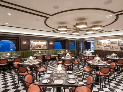 Blick in das L'Atelier Bistrot der MSC Grandiosa. Der elegante Restaurantbereich ist mit schwarz-weißem Schachbrettboden, orange-schwarzen Stühlen und einer langen olivgrünen Bank ausgestattet. Runde Fenster zeigen eine blaue Reflexion. Große goldene Kronleuchter hängen von der Decke, die mit braunen Holzleisten verziert ist. Tische sind eingedeckt, aber keine Personen sind sichtbar.