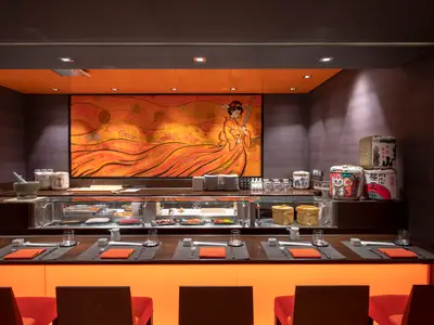 Die Kaito Sushi Bar an Bord der MSC Grandiosa. Eine Sushi-Theke mit Auslage und gedeckten Plätzen im Vordergrund. Ein großes, orangefarbenes japanisches Kunstwerk und Sake-Fässer schaffen ein stimmungsvolles Ambiente.