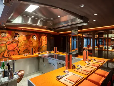 Innenansicht des Kaito Teppanyaki Restaurants der MSC Grandiosa. Es zeigt den Teppanyaki-Grill, orange Tische mit Gedecken und eine große Wandmalerei mit Geishas.
