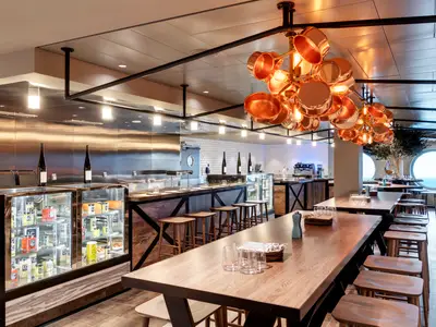 Modernes Hola! Tapas Restaurant an Bord der MSC Grandiosa. Es zeigt lange Holztische, eine Bar mit Hockern, runde Fenster mit Meerblick und dekorative Deckenlampen aus Kupfertöpfen.