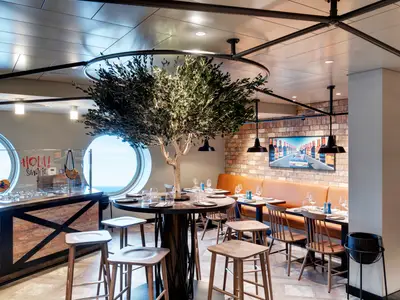 Die Hola! Tapas Bar an Bord der MSC Grandiosa. Ein großer Olivenbaum, hohe Tische, Ledersitzbänke an einer Ziegelwand und Bullaugen mit Meerblick.