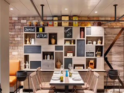 Einladende Hola! Tapas Bar by Ramon Freixa auf der MSC Grandiosa. Ein gedeckter Tisch steht vor einer Wand aus Holzwuerfeln mit Tafelbeschriftungen, Keramik und historischen Dosen.