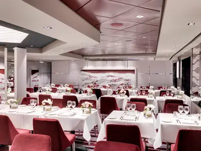 Innenansicht eines eleganten Restaurants an Bord der MSC Grandiosa, komplett mit weiß gedeckten Tischen, roten Stühlen und zeitgenössischer Kunst an den Wänden. Der Raum ist hell beleuchtet und ohne Gäste.