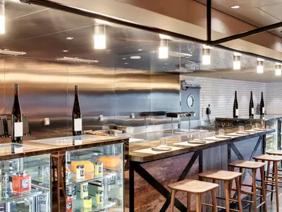 Moderne Hola! Tapas Bar der MSC Grandiosa: Holztheke mit Barhockern, Edelstahl, Weinflaschen und gedeckten Plaetzen. Links Kuehlvitrinen mit Kaese. Im Hintergrund weisse Ziegelwand und Schiffstür.