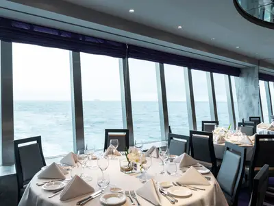 Elegantes Restaurant an Bord der MSC Grandiosa mit festlich gedeckten Tischen. Große Fensterfront bietet Panoramablick auf das Meer, ein Schiff in der Ferne.