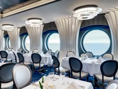 Ein weitläufiges, elegantes Restaurant an Bord der MSC Grandiosa mit zahlreichen gedeckten Tischen, modernen Lampen und runden Fenstern, die den Blick auf das offene Meer freigeben.