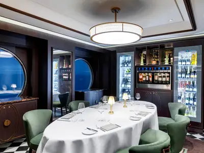 Ein elegantes Spezialitätenrestaurant, L'Atelier Bistrot, an Bord der MSC Grandiosa. Ein langer, weiss gedeckter Tisch mit grünen Stühlen, umgeben von dunklen Holzvertäfelungen, Weinregalen und einem Bullaugenfenster.