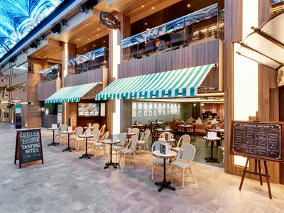 Das L'Atelier Bistrot an Bord der MSC Grandiosa. Aussenbestuhlung mit Tischen, Stühlen und gestreiften Markisen. Holzfassade, im Hintergrund Promenade mit blauem LED-Himmel. Keine Personen.