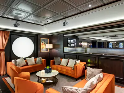 Lounge an Bord der MSC Grandiosa: orangefarbene Sofas, dunkle Holzvertäfelungen, rundes Lichtobjekt an der Wand. Bar hinter geriffelter Glaswand im Hintergrund.