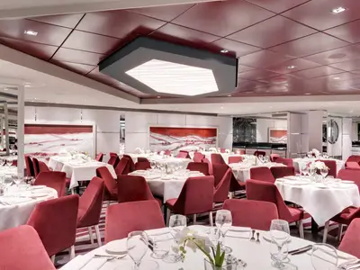Ein elegantes Restaurant an Bord der MSC Grandiosa. Zahlreiche Tische mit weissen Tischdecken und roten Samtstuehlen sind bereit. Die Waende zeigen rote Kunstwerke. Eine moderne Lichtinstallation dominiert die Decke.