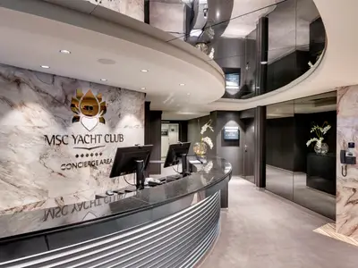 Der elegante Concierge-Bereich des MSC Yacht Clubs an Bord der MSC Grandiosa, mit einer Marmorwand, goldenem Logo, schwarzem Schreibtisch und Spiegeldecken.