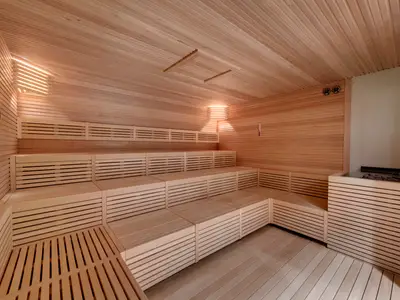 Eine helle, moderne Holzsauna an Bord der MSC Grandiosa. Sie ist mit mehrstufigen Baenken aus hellem Holz ausgestattet und wird durch indirekte Beleuchtung warm in Szene gesetzt. Rechts ist ein Saunaofen sichtbar.
