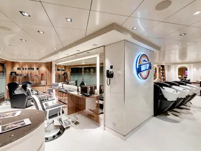 Ein stilvoller Barbershop an Bord der MSC Grandiosa mit mehreren Friseurstühlen und hellen Waschbecken. Produkte sind in Regalen ausgestellt, ein beleuchtetes Logo ist sichtbar.