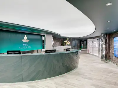 Der moderne Empfang des MSC Aurea Spa auf der MSC Grandiosa. Eine geschwungene Theke, dahinter eine aqua-farbene Wand mit dem Spa-Logo (Meditationsfigur). Ambiente ist ruhig und entspannend.
