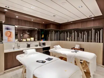 Zwei weiß bezogene Massageliegen in einem modernen Wellnessraum an Bord der MSC Grandiosa. Dunkles Holz und helle Wände prägen den Raum. Links eine Rezeption mit Waschbecken und Pflegeprodukten.