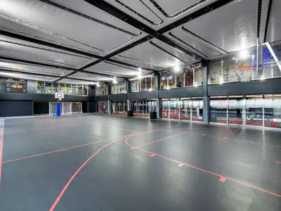 Ein moderner, dunkler Indoor-Sportplatz an Bord der MSC Grandiosa. Rote Linien markieren Spielfelder für Basketball und Fußball. Links sind ein Basketballkorb, zwei Fußballtore und eine digitale Anzeigetafel zu sehen. Rechts steht ein weiterer Basketballkorb. Die Decke ist hell und mit Beleuchtungselementen versehen. Im Hintergrund befinden sich Fensterfronten zu weiteren Bereichen des Schiffs.