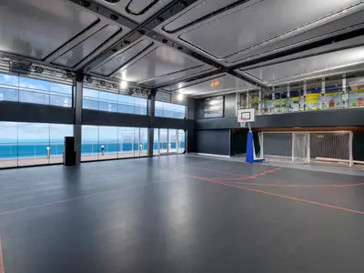 Ein moderner Indoor-Sportplex an Bord der MSC Grandiosa. Der graue Sportboden mit roten Markierungen bietet Platz für Basketball und Fußball. Ein Basketballkorb und ein Fußballtor stehen bereit. Große Fensterfronten geben den Blick auf das weite, blaue Meer und ein Außendeck frei. Über dem Tor sind durch weitere Fenster bunte Dekorationen eines Kinderbereichs zu sehen. Die Decke ist mit Lichtinstallationen versehen, und ein Scoreboard hängt an der Wand.