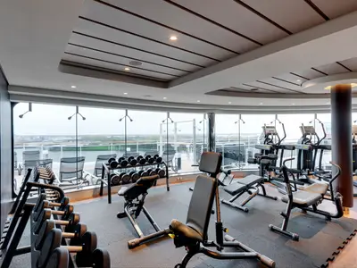 Ein moderner Fitnessraum an Bord der MSC Grandiosa mit Technogym-Geräten, Hanteln und Bänken, umgeben von großen Fenstern mit Blick auf das Meer und die Hafenlandschaft. Der Raum ist hell beleuchtet und mit dunklen Sportmatten ausgelegt.