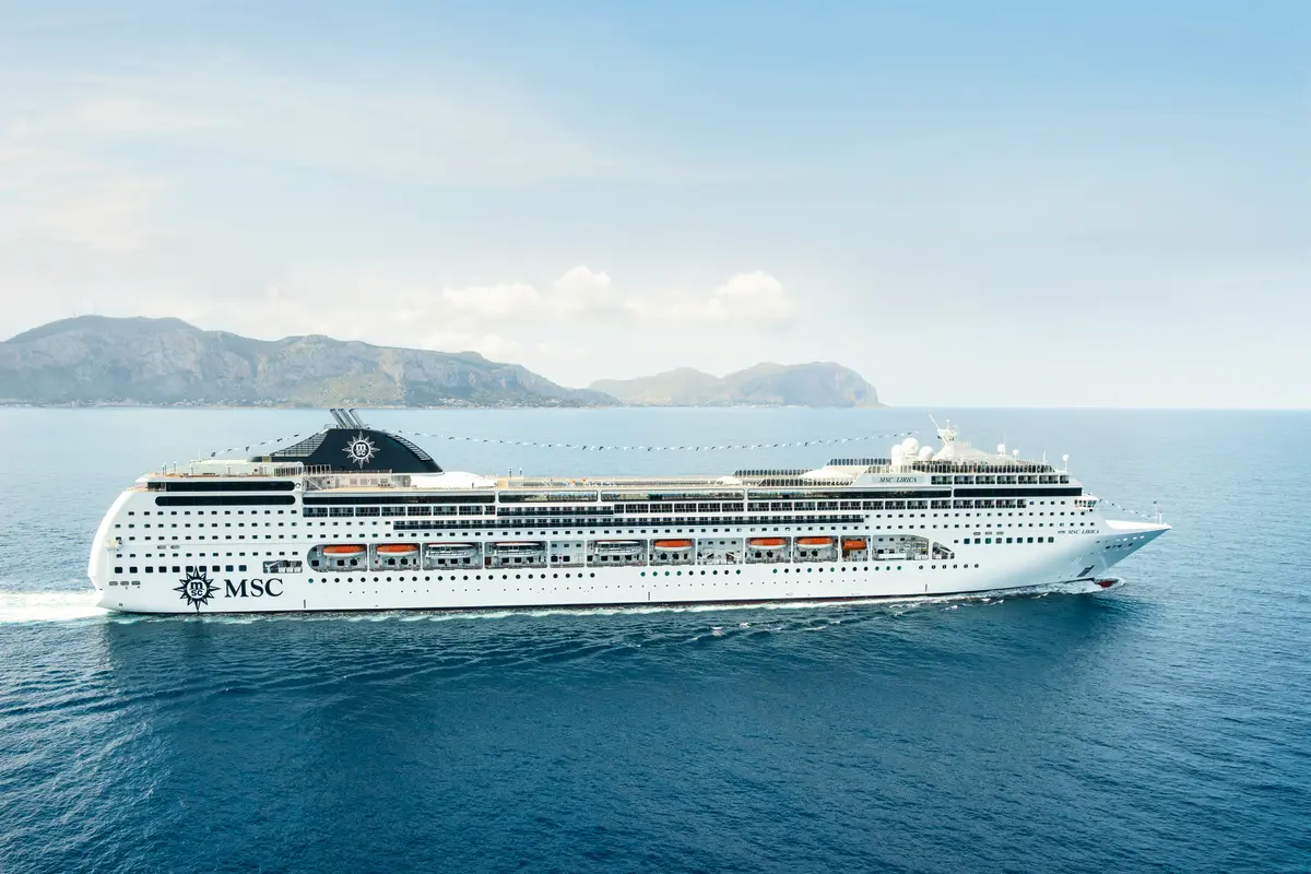 Die MSC Lirica, ein weißes Kreuzfahrtschiff mit dem MSC-Logo am Heck und Schornstein, fährt auf blauem Meer vor einer felsigen Küste und Bergen unter einem leicht bewölkten Himmel.