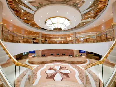 Elegantes Atrium der MSC Lirica mit Rezeption, geschwungenen Treppen und Goldgeländern. Marmorboden und eine zentrale Deckenleuchte dominieren den mehrstöckigen Raum.