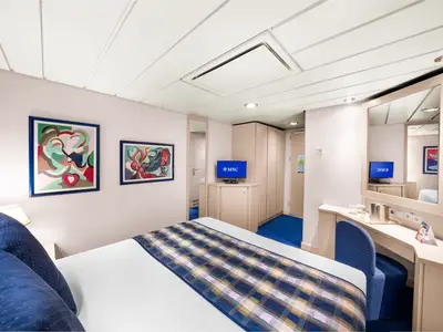 Helle Aussenkabine an Bord der MSC Lirica. Das Doppelbett hat blaue Kissen. Ein Schreibtisch mit Spiegel und TV ist zu sehen. Moderne Kunstwerke und ein blauer Teppichboden ergänzen die helle Einrichtung.