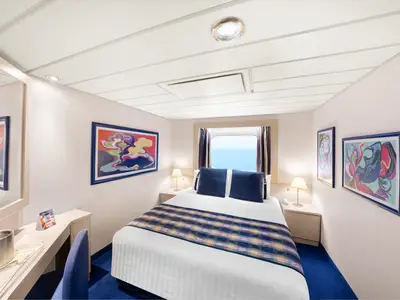 Helle Aussenkabine an Bord der MSC Lirica mit Doppelbett, blauem Teppich und Fenster mit Meerblick. Links ist ein Schreibtisch zu sehen. Die Wände sind mit abstrakter Kunst dekoriert.