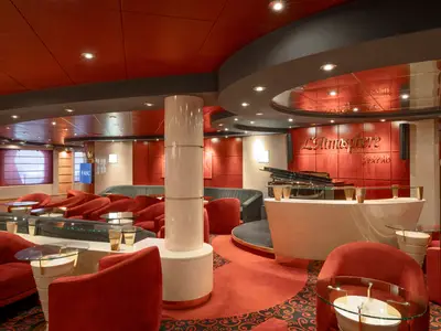 Eine Weitwinkelaufnahme der L'Atmosphere Piano Bar an Bord der MSC Lirica, mit roten Armsesseln, Couchen, glänzenden Tischen und einem Flügel. Der Raum ist in Rottönen gehalten mit einer Bühne.