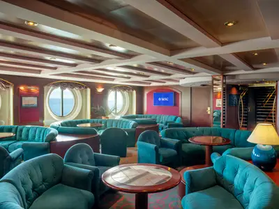 Einladende Lord Nelson Pub Bar & Lounge an Bord der MSC Lirica mit teal-farbenen Samtsofas, Sesseln, Holztischen und runden Fenster mit Meerblick. Ein Bildschirm zeigt das MSC Logo.