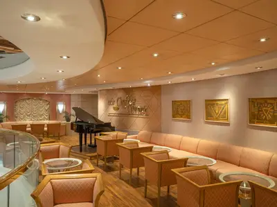 The Coffee Corner Lounge der MSC Lirica: beigefarbene Sofas, Sessel, Tische, schwarzer Flügel, goldene Wandkunst. Warme, einladende Atmosphäre.