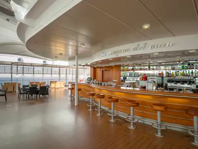 Die helle Bar 'La Canzone Del Mare' an Bord der MSC Lirica mit eleganter Holztheke, Barhockern und Spirituosenregalen. Ein überdachter Aussenbereich bietet Tische und Meerblick.