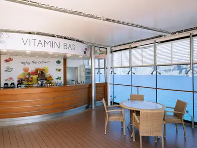Die helle Vitamin Bar an Bord der MSC Lirica mit Holztresen, bunten Fruchtdisplays und Rattanmöbeln. Große Fensterfront bietet Panoramablick auf das blaue Meer und eine felsige Küste. Eine entspannte Atmosphäre lädt zum Verweilen ein.