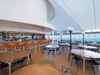 Ein heller, offener Bar- und Restaurantbereich an Bord der MSC Lirica mit Holztheke, vielen Tischen und Meerblick unter einer weissen Zeltüberdachung.