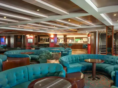 Der Lord Nelson Pub an Bord der MSC Lirica zeigt einen eleganten Bereich mit zahlreichen türkisfarbenen Samtsofas, runden Holztischen, Pflanzen und einer Bar im Hintergrund. Das Licht ist warm und einladend.