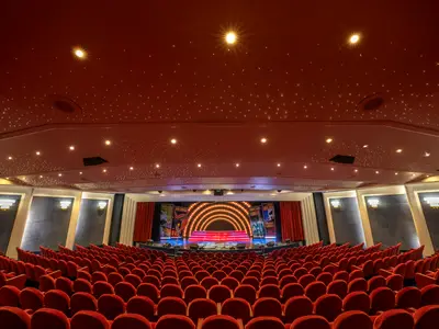 Blick in das The Broadway Theatre an Bord der MSC Lirica. Rote Samtsitze säumen den Gang zur Bühne. Die Bühne ist hell beleuchtet mit einem halbkreisförmigen Lichtbogen und Plakaten an den Seiten.