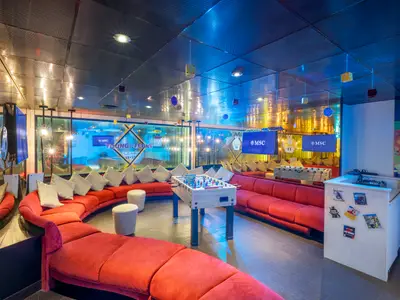 Ein lebhafter Young & Teens Club an Bord der MSC Lirica mit einer grossen roten Sofalandschaft, weissen Kissen und einem Kickertisch in der Mitte. Mehrere Bildschirme zeigen das MSC Logo. Die Decke reflektiert das Licht.
