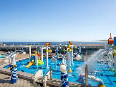 Ein farbenfroher Doremi Spray Park an Bord der MSC Lirica auf dem Oberdeck mit diversen Wasserspielen, Fontänen, einer gelben Rutsche und Blick auf das blaue Meer. Viel Wasser spritzt im Bereich.