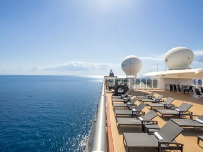 Weitwinkelaufnahme des sonnigen Außendecks der MSC Lirica mit zahlreichen leeren Liegestühlen. Im Hintergrund sind das weite Meer und der blaue Himmel sichtbar, mit Satellitenschüsseln auf dem oberen Deck.