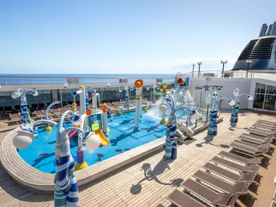 Doremi Spray Park der MSC Lirica: Bunter Wasserspielplatz mit Rutschen und Fontänen auf dem sonnigen Oberdeck. Im Hintergrund weites Meer und Schiffsschornstein. Holzdeck mit Liegestühlen.