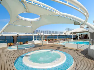 Das sonnige Deck der MSC Lirica zeigt zwei sprudelnde Whirlpools und einen Pool. Ein modernes weißes Sonnensegel spendet Schatten, während die Weite des Meeres zu sehen ist. Entspannung pur an Bord.