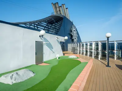Grüner Mini-Golf-Parcours mit Felsen auf dem Deck der MSC Lirica. Heller Himmel, dunkler Schornstein mit MSC-Logo, Balustrade und das offene Meer sind im Hintergrund sichtbar.