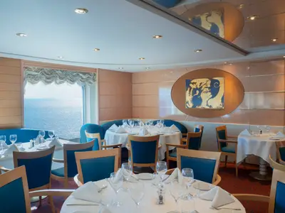 Ein elegantes Restaurant an Bord der MSC Lirica mit Tischen, blauen Stühlen und Bänken. Ein großes Fenster bietet einen weiten Blick auf das glitzernde Meer.