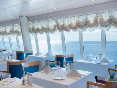 Restaurant L'Ippocampo auf der MSC Lirica. Elegant gedeckte Tische, blaue Stühle. Große Fenster mit Raffrollos geben den Blick auf das weite, glitzernde Meer frei.