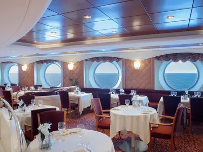 Ein Blick in das stilvolle La Bussola Restaurant an Bord der MSC Lirica. Zahlreiche Tische mit weissen Decken und roten Stühlen, umrahmt von grossen Bullaugen mit Meerblick.