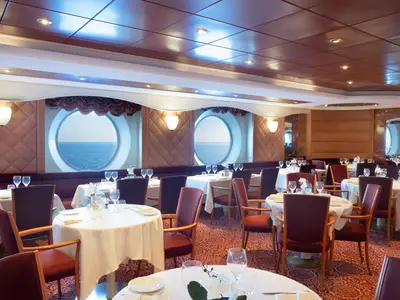 Ein elegantes Restaurant an Bord der MSC Lirica, "La Bussola", mit vielen gedeckten Tischen, roten Stühlen und großen runden Bullaugen, die den Blick auf das offene Meer freigeben.
