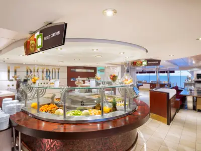 Grosses "Fruit Station" Obstbuffet im "Le Bistrot" der MSC Lirica. Frisches Obst, Salate und Teller. Sitzbereiche und Meerblick durch Panoramafenster sind zu sehen.