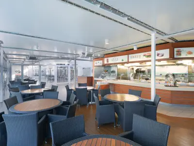 Heller, weitläufiger Buffetbereich 'Hamburger Paradise' an Bord der MSC Lirica. Zahlreiche Tische und dunkle Stühle aus Geflecht sind zu sehen. Im Hintergrund eine lange Theke mit Speisen und grossen Bildern von Hamburgern, Pommes und Zwiebeln. Tageslicht durchflutet den Raum.