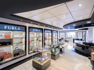 Ein moderner, gut beleuchteter Luxus-Shop mit Designer-Handtaschen von Furla und Coccinelle an Bord der MSC Lirica. Glasvitrinen und ein geschwungener Tresen praesentieren die Waren.