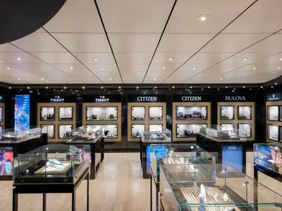 Ansicht eines luxuriösen Uhren- und Schmuckgeschäfts an Bord der MSC Lirica. Mehrere Vitrinen präsentieren Uhren von Marken wie Balmain, Tissot, Citizen und Bulova. Eine grüne Halskette liegt in einer Vitrine im Vordergrund.