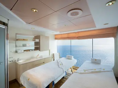 Ein heller MSC Aurea Spa-Behandlungsraum an Bord der MSC Lirica mit zwei bezogenen Massagebetten und bodentiefen Fenstern, die einen weiten Blick auf das glitzernde blaue Meer bieten. Links ein Waschtisch und ein Duschbereich.