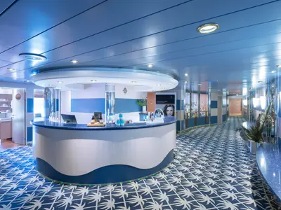 Heller Empfangsbereich des MSC Aurea Spa auf der MSC Lirica, gestaltet in blau-weiß. Ein geschwungener Tresen zeigt Pflegeprodukte. Rechts führt ein Flur mit Vitrinen entlang. Der Boden hat ein blaues Palmenmuster.